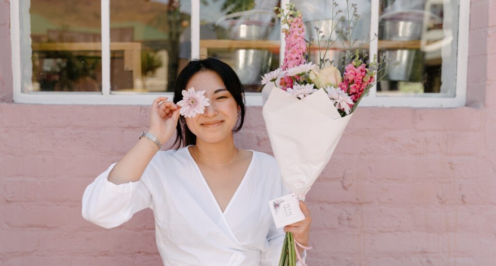 A woman smiles holding a pink posy in one hand and a chrysanthemum bud over her eye in the other.